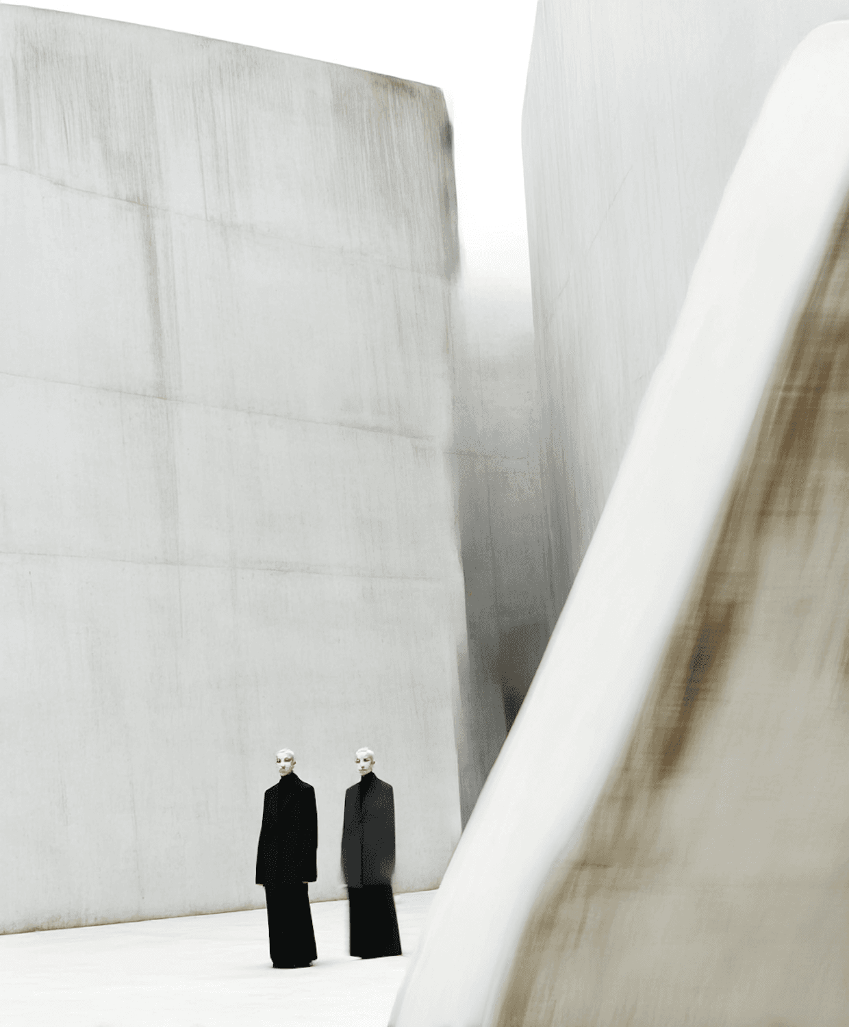Duas figuras vestidas de preto diante de grandes paredes de concreto claro